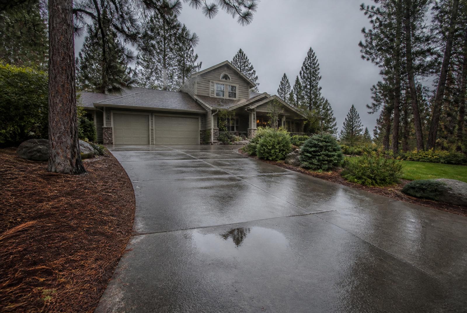 A Spokane residential driveway after spring rain with a small puddle in a subtle low spot, surrounded by pine trees and an Inland Northwest home under overcast skies